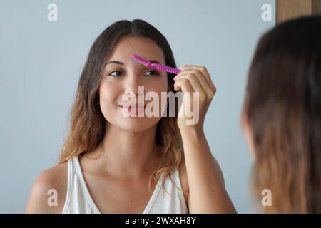 Dermaplaning. Bella ragazza ispanica che si radica le sopracciglia con un rasoio sullo specchio di casa. Rimozione dei peli del viso. Foto Stock