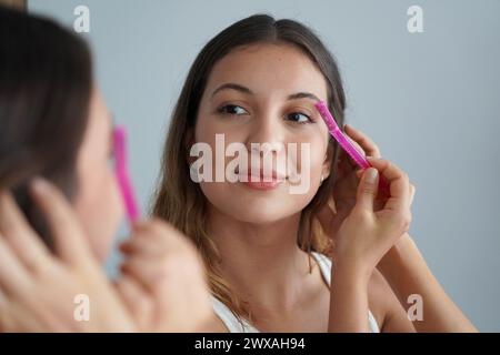 Dermaplaning. Primo piano di una bella ragazza che si rasa le sopracciglia con il rasoio allo specchio di casa. Rimozione dei peli del viso. Foto Stock