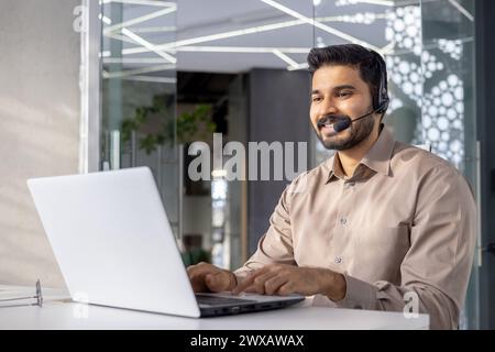 Un uomo professionista con una cuffia che utilizza un computer portatile in un ufficio moderno, che trasmette assistenza clienti e tecnologia. Foto Stock