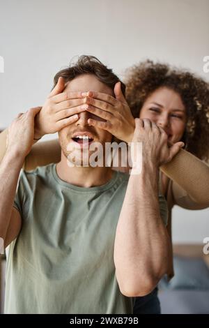 Coppia adorante, giovane donna riccia che copre gli occhi di un uomo bruno, che condivide un momento amorevole con i sorrisi Foto Stock