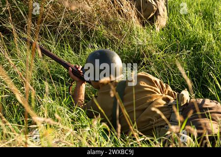 Ricostruzione storica. Un soldato di fanteria americano della seconda guerra mondiale spara nascosto nell'erba alta. Vista dal retro Foto Stock