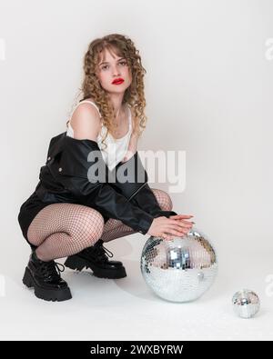 Bella ragazza adulta che tiene in mano una sfera da discoteca a specchio e guarda la macchina fotografica. Giovane donna gen z accovacciata su sfondo bianco. Donna seria Foto Stock