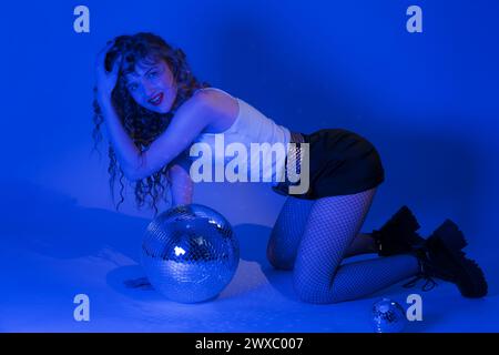 Donna sensuale con ballo a specchio che si posa in ginocchio in un'illuminazione blu al neon durante la festa di festa nel nightclub. Giovane donna Foto Stock