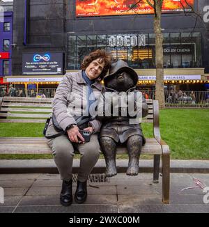 Una turista donna seduta su una panchina accanto a una statua di bronzo dell'Orso Paddington, dietro di loro ODEON Luxe è uno dei cinema più iconici al mondo. Foto Stock