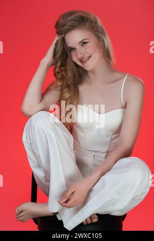 Ritratto di una giovane donna bionda sorridente, seduta sullo sgabello da bar con i piedi in alto e con lo sguardo lontano. Immagine di una giovane modella di moda femminile Foto Stock