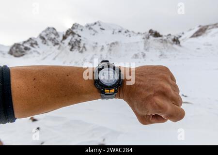 Mano dell'uomo con orologio elettronico che mostra l'altitudine con montagne innevate sullo sfondo. Concetto di arrampicata ed escursionismo. Foto Stock