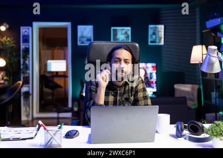 Ritratto di un giovane uomo seduto di fronte al suo portatile alla sua scrivania di casa, che lavora da casa. Un uomo serio che tiene in mano una penna mentre guarda la fotocamera, si prepara al lavoro e alla ricerca. Foto Stock