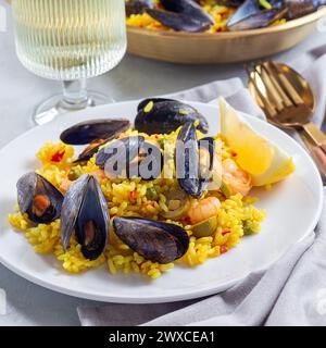 Paella di pesce con cozze e gamberi, su un piatto bianco, servita con limone e vino bianco, formato quadrato Foto Stock