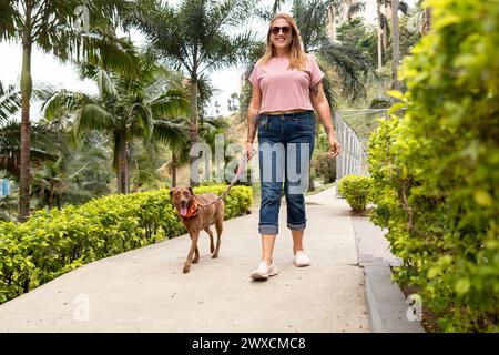 Una donna di mezza età sorride e guarda la telecamera mentre cammina con il suo cane, che conduce al guinzaglio attraverso un parco. Foto Stock