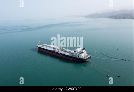 Ripresa aerea di una massiccia petroliera ancorata al largo vicino a un paesaggio urbano costiero in una giornata limpida. Foto Stock