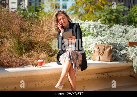 Donna d'affari sorridente con caffè seduto a parlare con uno smartphone in mano all'aperto. Foto Stock