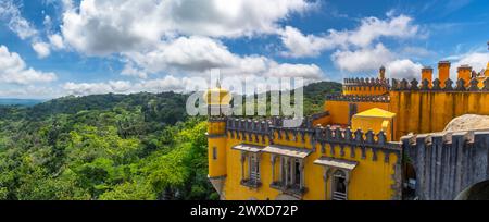 Ampio angolo di veduta esterna della sommità della torre di guardia gialla e dell'attico del Palácio da pena, sotto un cielo azzurro nuvoloso e soleggiato, con il frondoso Monte Sintra Foto Stock