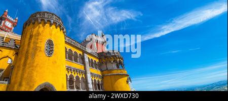 Ampia vista esterna del Palácio da pena giallo e rosso con il suo cortile ad arco, sotto un cielo azzurro soleggiato con le montagne all'orizzonte. Sintra Foto Stock