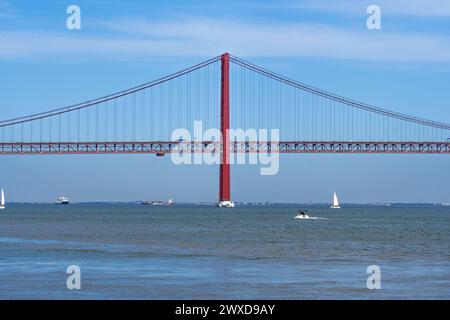 Nave portacontainer, motoscafi e barche a vela che navigano sul fiume Tago attraversando il ponte sospeso in acciaio rosso 25 de Abril con traffico stradale per l'accesso Foto Stock