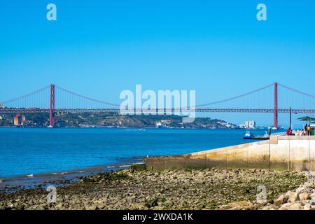 Vista panoramica del ponte sospeso in metallo rosso 25 de Abril di Lisbona, come il Golden Gate, dalla costa della spiaggia con persone sedute in un bar Foto Stock