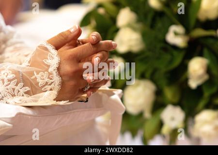 le mani di una sposa, adornate da un delicato anello nuziale, sono piegate in preghiera sopra il suo abito in pizzo, con rose bianche sullo sfondo Foto Stock