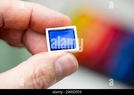 Una mano che mostra un piccolo cubo di acquerello blu con uno sfondo multicolore. Formato orizzontale. Scegliere, scegliere, selezionare Foto Stock