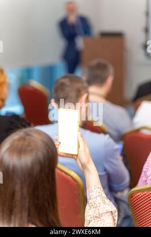 Una donna scatta una conferenza con la fotocamera di uno smartphone durante una conferenza o un seminario di lavoro. Il pubblico ascolta il docente alla conferenza nell'auditorium. Foto Stock