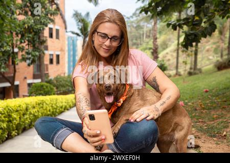 Donna seduta con gli occhiali da lettura che si fa un selfie con il suo animale domestico seduto nel parco. Foto Stock