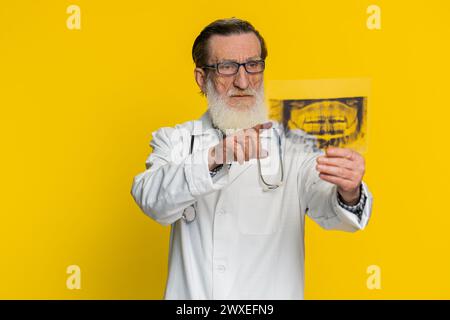 Anziano medico anziano ortodontista esamina un'immagine radiografica panoramica dei denti della mascella. Modello 3D della bocca del paziente, scansione RM. Odontoiatria orale. Nonno di Stomatologia su sfondo giallo Foto Stock