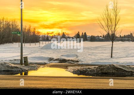 Colore arancione riflesso sulla pozzanghera dalla neve sciolta ai margini del marciapiede al tramonto all'inizio della stagione primaverile Foto Stock