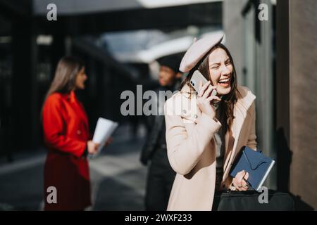 La giovane donna alla moda ama una conversazione allegra sul suo smartphone, ridendo mentre cammina accanto ai colleghi concentrati all'aperto. Foto Stock