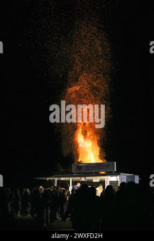 Sythen, NRW, Germania. 30 marzo 2024. Il fuoco di Pasqua è visibile dietro un banco di ristoro. La gente del posto e i visitatori si riuniscono per l'annuale falò di Pasqua presso la fattoria Prickingshof di Sythen, Vestfalia (Münsterland). L'illuminazione dei falò di Pasqua è stata una tradizione per molti secoli in Germania, di solito il sabato Santo o la domenica di Pasqua. I vigili del fuoco e i servizi di emergenza garantiscono che il fuoco sia tenuto al sicuro mentre i volontari hanno ispezionato attentamente la legna da ardere per assicurarsi che nessuna fauna selvatica si sia rifugiata nei rami. Crediti: Imageplotter/Alamy Live News Foto Stock