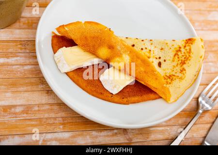 Al centro dello shot c'è un piatto con un'appetitosa colazione - morbidi pancake ruvide con pezzi di formaggio brie cremoso. Il piatto è completato da una tazza di caldo Foto Stock