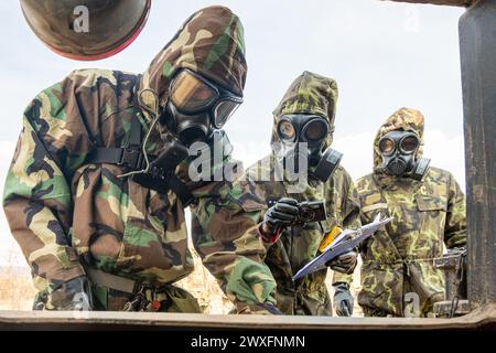 Tre uomini in tuta di nocciola guardano da una finestra. Indossano maschere antigas e sembrano trovarsi in un ambiente militare Foto Stock