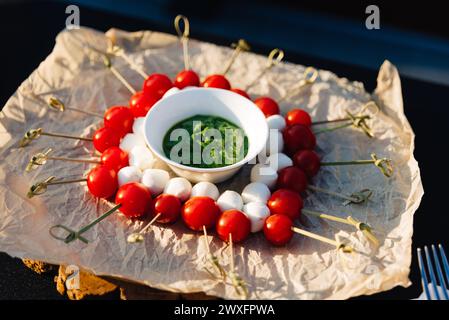 Una deliziosa disposizione di spiedini caprese, con pomodori ciliegini, palline di mozzarella e un vibrante pesto di basilico su carta naturale. Foto Stock