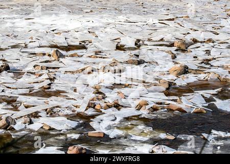 Un fiume poco profondo coperto da frammenti di ghiaccio rotto sostenuto da fango e roccia. Un po' d'acqua scorre tra il ghiaccio. Foto Stock