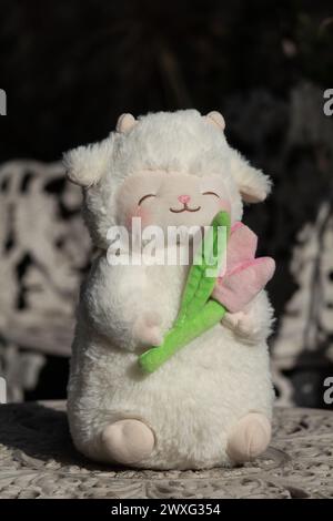 Simpatico peluche soffice di agnello bianco che arrossisce e tiene in mano un tulipano rosa seduto sopra un tavolo di ceramica bianca Foto Stock