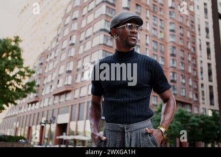 Il bell'elegante uomo afroamericano si erge in strada con un abbigliamento alla moda, indossando un berretto, occhiali da sole e una t-shirt nera, moderna moda maschile Foto Stock