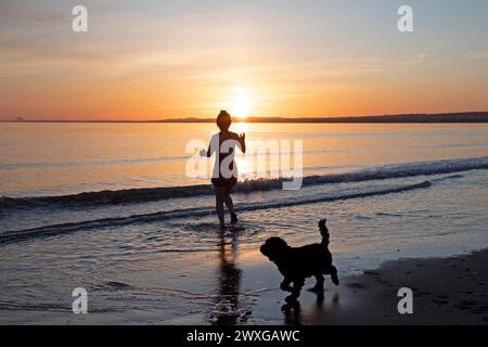 Portobello, Edimburgo, Scozia, Regno Unito. 31 marzo 2024. Splendida alba per il benvenuto nel periodo estivo britannico sul mare scozzese, così come la domenica di Pasqua. Nella foto: Makiko Konishi fa un tuffo fresco nel Firth of Forth all'alba mentre il suo cockapoodle Renzo aspetta sulla riva, temperatura di 6 gradi centigradi. Credito: Arch White/alamy notizie dal vivo. Foto Stock