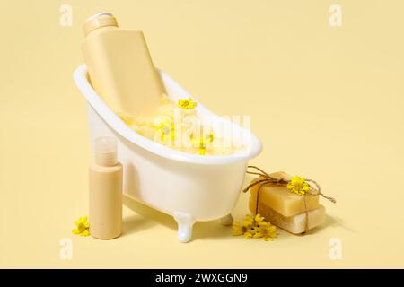 Composizione con mini vasca da bagno, bottiglie di prodotti cosmetici e fiori su sfondo giallo Foto Stock