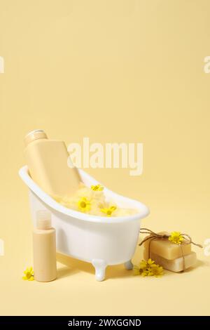 Composizione con mini vasca da bagno, bottiglie di prodotti cosmetici e fiori su sfondo giallo Foto Stock