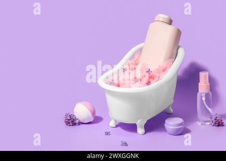 Composizione con mini vasca da bagno, bottiglie di prodotti cosmetici e bomba su sfondo lilla Foto Stock