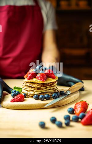 Uomo che serve pancake panna su un tavolo di legno con fragole e mirtilli, luce calda, bar Foto Stock