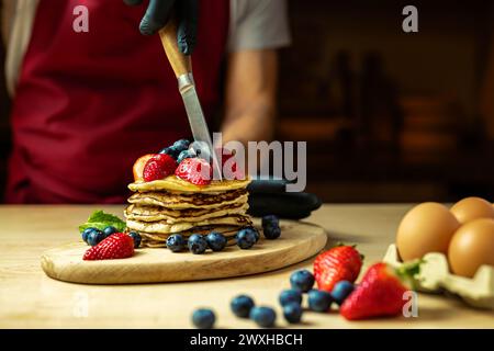 uomo che taglia pancake con panna, fragole e mirtilli su un asse di legno, luce calda, bar. Foto Stock