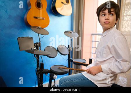 Sicuro batterista caucasico adolescente musicista seduto alla batteria in uno studio di musica retrò a casa, tenendo la bacchetta e guardando da parte sognando. Elettr Foto Stock