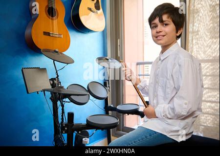 Giovane batterista in camicia bianca e jeans blu, seduto al drum set nel suo studio di musica retrò, tenendo la bacchetta e sorridente Foto Stock