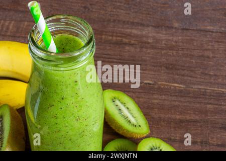 Frullato verde a base di kiwi e banana in una bottiglia di vetro con frutta fresca su fondo rustico in legno. Sana bevanda disintossicante estiva Foto Stock