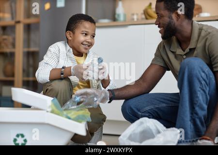 Ritratto di un ragazzo afroamericano sorridente che ordina la plastica per il riciclaggio a casa con il padre Foto Stock