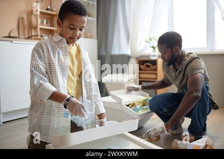 Ritratto di un giovane ragazzo nero che smista i rifiuti a casa con il padre e mette la bottiglia di plastica nello spazio di copia del cestino di riciclaggio Foto Stock