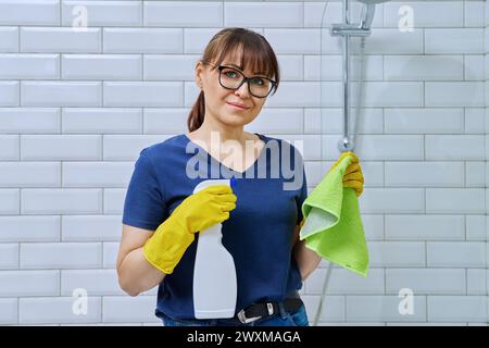 Donna che indossa guanti con un panno detergente spray che esegue la pulizia in bagno Foto Stock