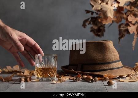 Whisky in vetro di cristallo su un tavolo di pietra grigia con foglie di quercia essiccate e cappello da uomo. Foto Stock