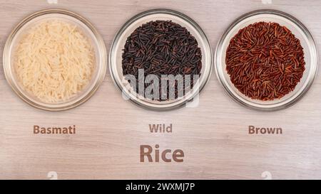 Tre ciotole trasparenti mostrano diverse varietà di riso - Basmati, Wild e Brown - ben etichettate, appoggiate su una superficie liscia in legno. L'assortimento Foto Stock