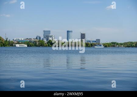 Vista dello skyline di Bucarest dall'altro lato di un tranquillo lago Herastrau con moderni edifici alti e lussureggiante vegetazione sullo sfondo in Romania Foto Stock