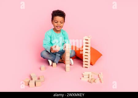 Foto a tutto il corpo di un bambino carino che gioca a cubi di blocchi di legno vestiti eleganti abiti ciano isolati su sfondo di colore rosa Foto Stock