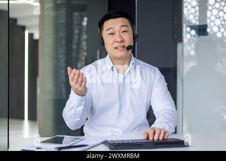 Uomo asiatico professionista in un ambiente di lavoro che effettua una chiamata con una cuffia, coinvolgendo la fotocamera. Foto Stock